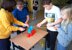 Grupa uczniów unosi kubeczek za pomocą sznurków. Wspólnie budują wieżę z kubków.